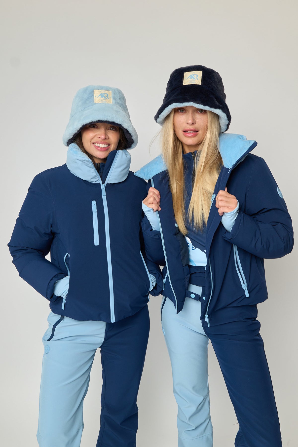 Two women modeling matching ski outfits from the brand Ruanua, featuring navy blue jackets with light blue accents, light blue pants, and winter hats, posed against a plain light gray studio background.