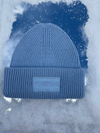 A blue ribbed knit beanie hat with a cuffed brim and a rectangular patch reading 'ruanua' on the front, set against a stylized icy blue background.