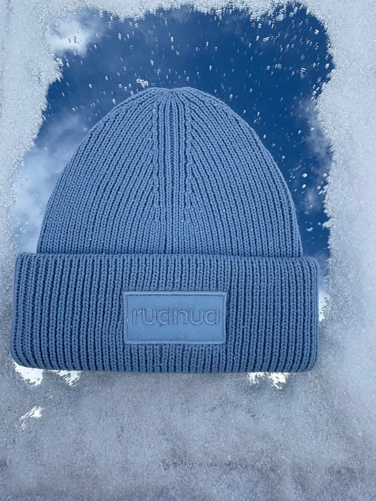 A blue ribbed knit beanie hat with a cuffed brim and a rectangular patch reading 'ruanua' on the front, set against a stylized icy blue background.