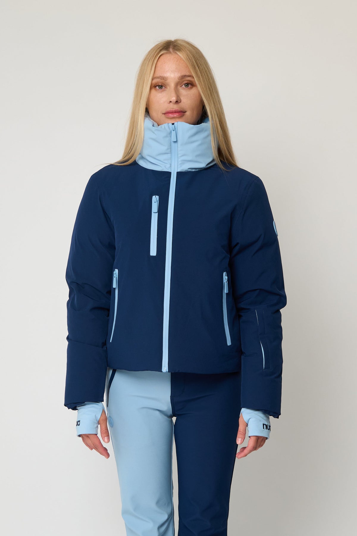 A woman modeling a ski jacket and matching ski pants in a studio setting. The jacket is navy blue with light blue accents, and the pants are split between light blue and navy, with light blue cuffs and hem.