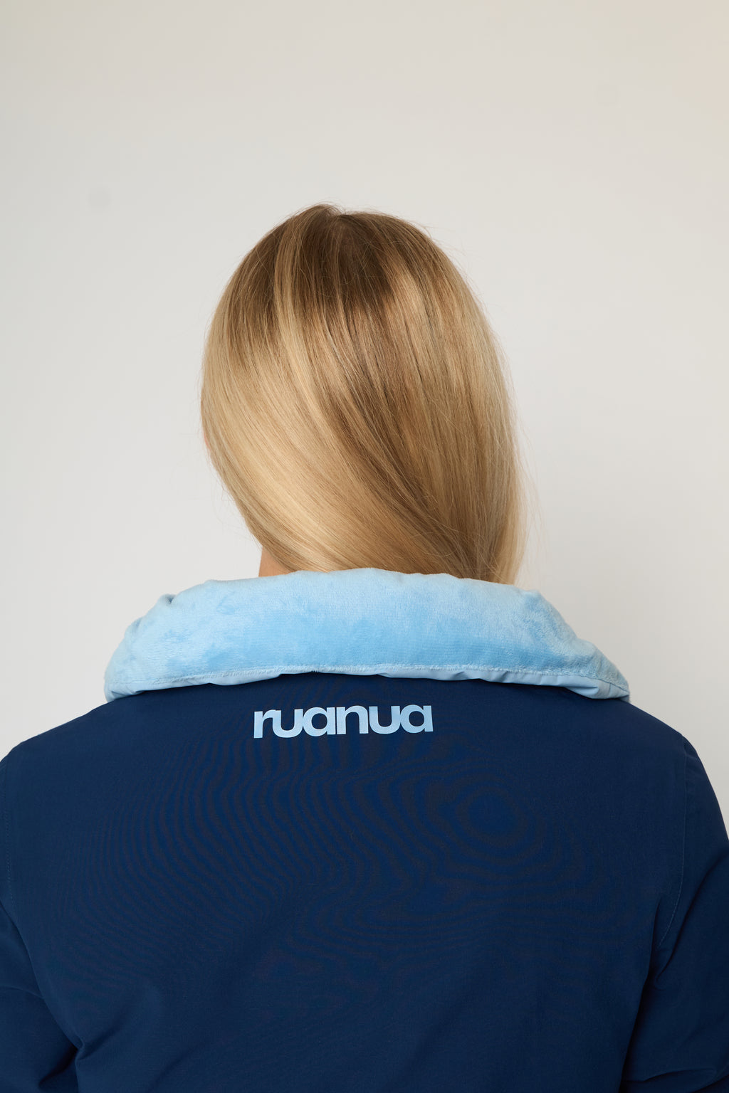 Back view of a person wearing a navy blue puffer jacket with a light blue fleece collar. The brand name 'ruanua' is printed in white on the upper back.