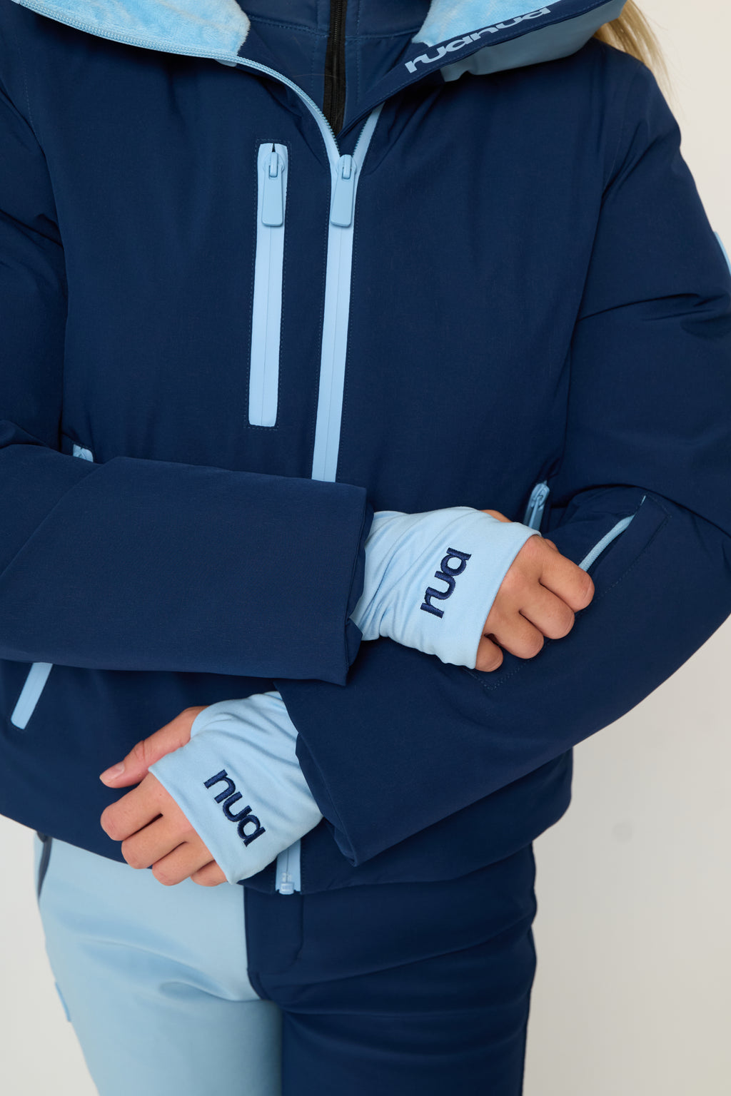 A close-up of a person wearing a navy ski jacket with light blue accents and matching light blue ski pants. The jacket has a front zipper, high neck funnel and light blue wrist warmers with the brand name 'rua' and 'nua' printed on them.