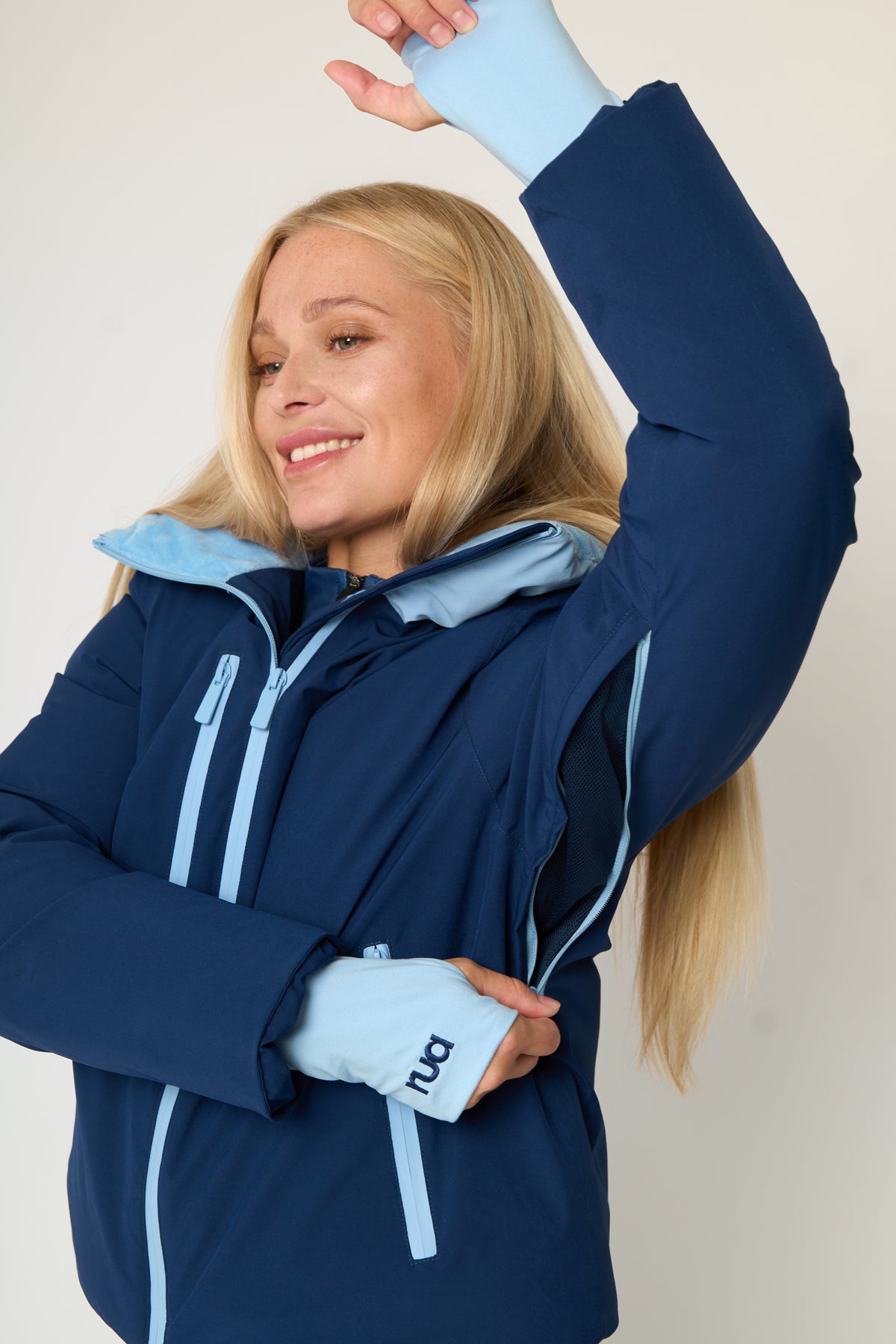 A woman wearing a navy blue ski jacket with light blue accents, including the high neck funnel lining, zipper trims, and storm cuffs, posing with one arm raised against a plain light background is unzipping her underarm air vents