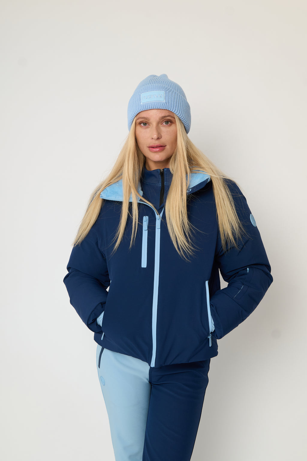 A blonde woman wearing a navy blue ski jacket with light blue accents, a matching light blue beanie, and light blue ski pants, standing against a plain off-white background.
