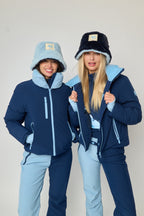 Two women modeling matching ski outfits from the brand Ruanua, featuring navy blue jackets with light blue accents, light blue pants, and winter hats, posed against a plain light gray studio background.