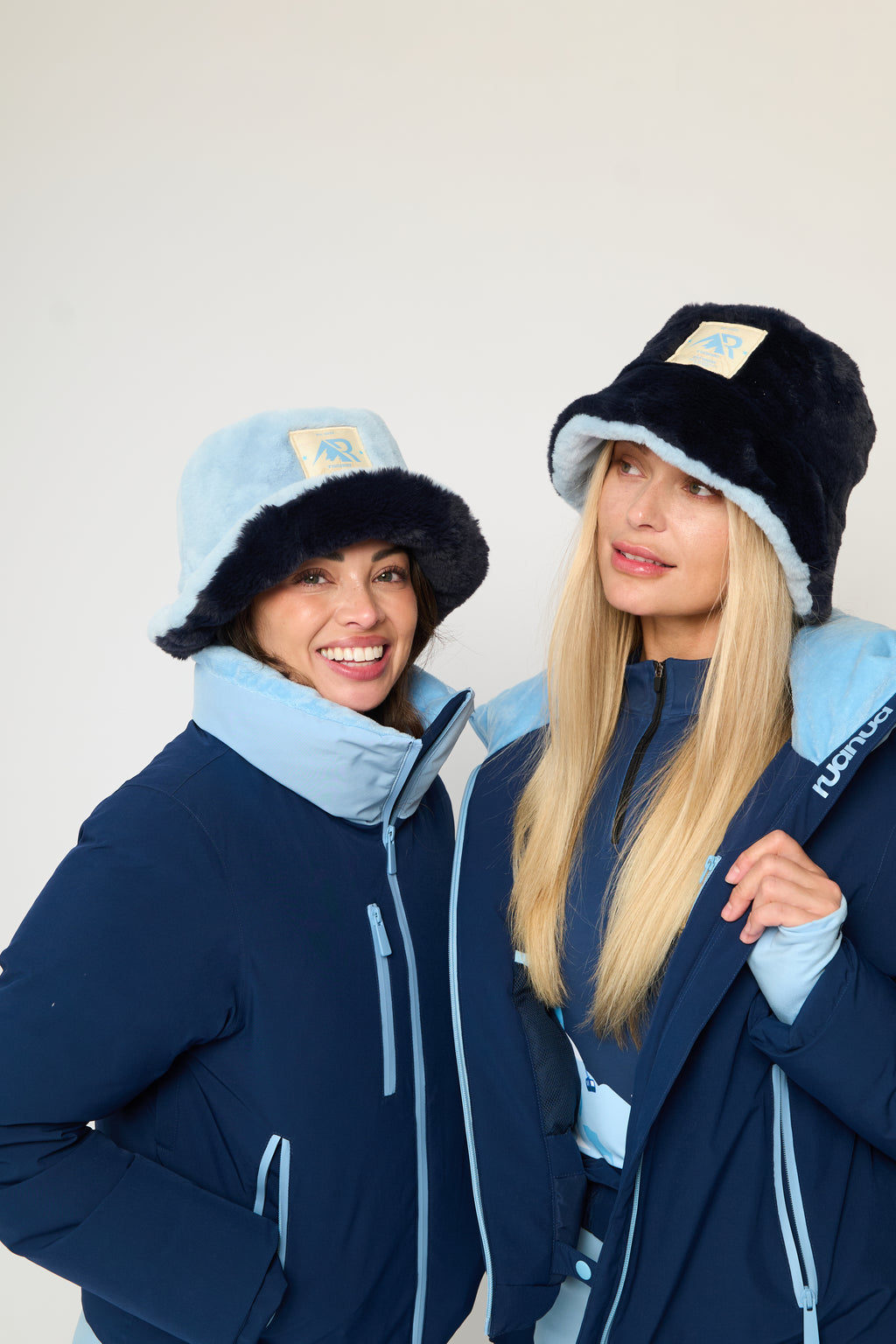 Two women modeling matching navy blue ski jackets with light blue accents and fuzzy bucket hats, posed against a plain off-white studio background.