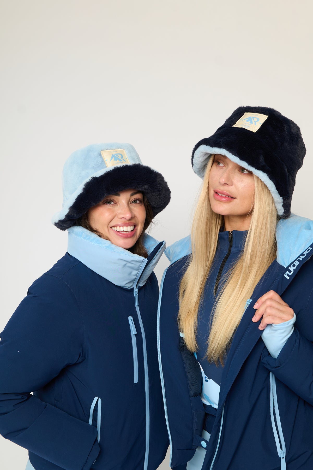 Two women modeling matching navy blue ski jackets with light blue accents and fuzzy bucket hats, posed against a plain off-white studio background.