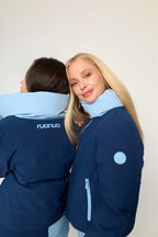 Two models wearing navy blue puffer jackets with light blue accents, including a high collar and a circular patch on the sleeve, against a plain off-white background.