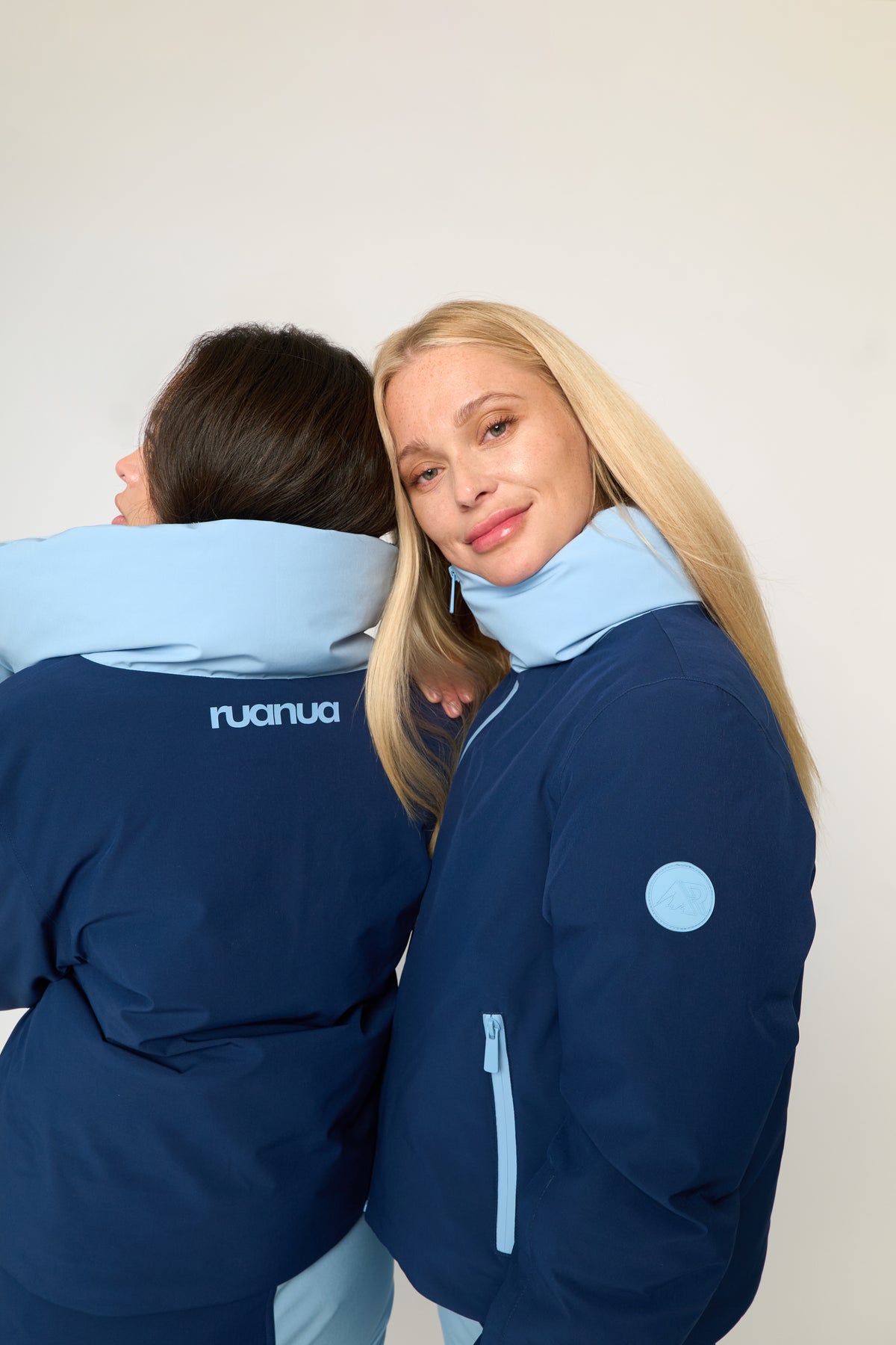 Two models wearing navy blue puffer jackets with light blue accents, including a high collar and a circular patch on the sleeve, against a plain off-white background.