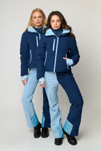Two women modeling matching ski outfits consisting of navy blue jackets with light blue accents and light blue pants, standing against a light gray studio background.