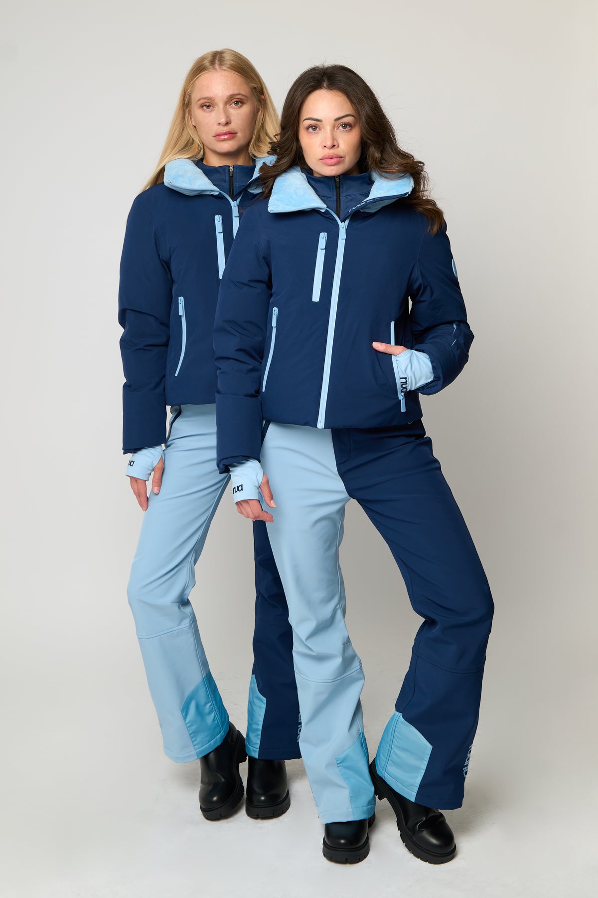 Two women modeling matching ski outfits consisting of navy blue jackets with light blue accents and light blue pants, standing against a light gray studio background.