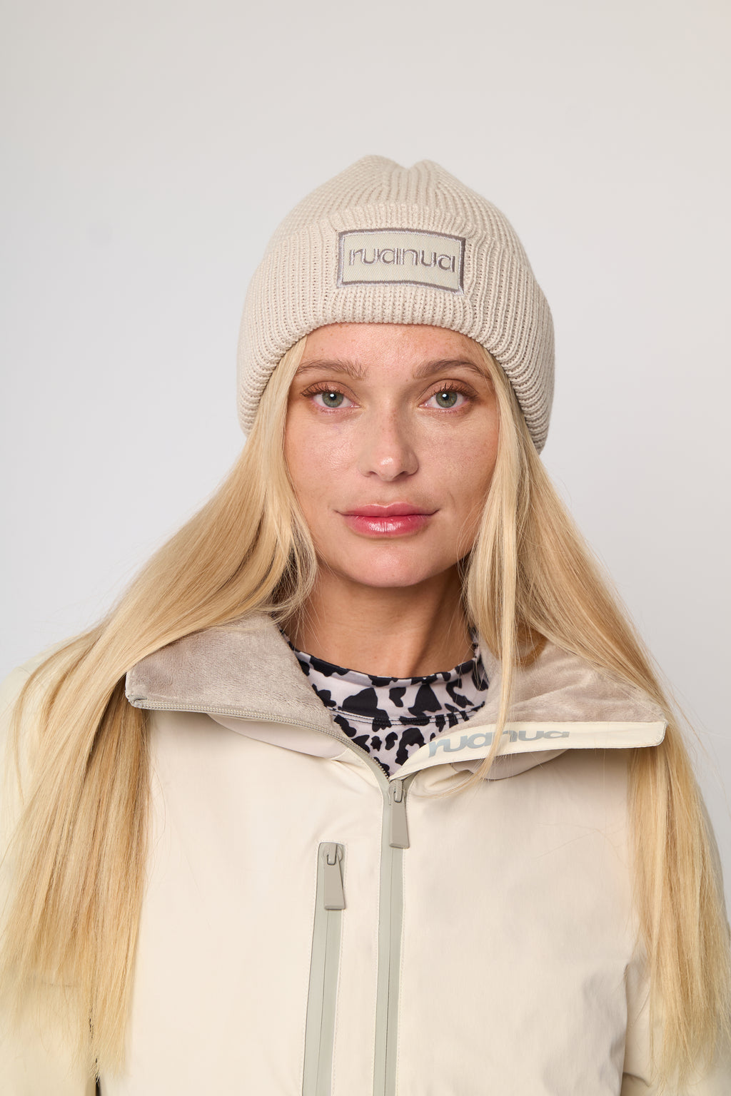 A woman with long blonde hair wearing a beige ribbed knit beanie with a rectangular logo patch that reads 'ruanua' and a cream-colored ski jacket with a soft fleece collar, photographed against a light gray studio background.