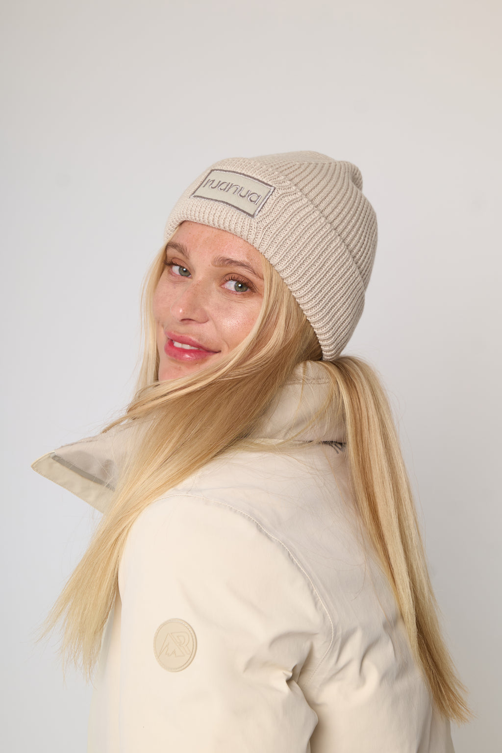A woman with long blonde hair wearing a beige ribbed knit beanie with a visible 'ruanua' brand patch and a cream-colored puffer jacket, photographed against a plain light gray background.