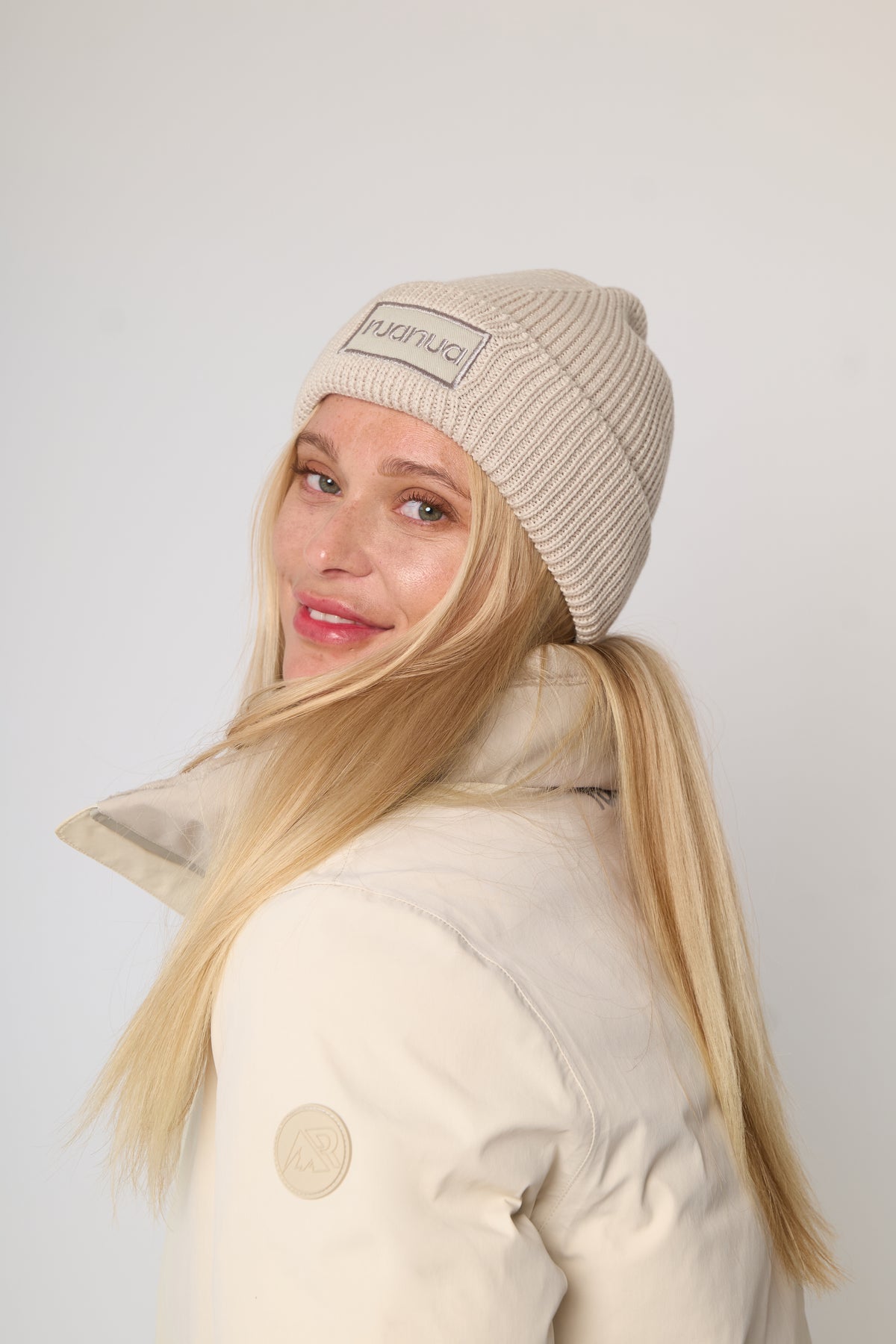 A woman with long blonde hair wearing a beige ribbed knit beanie with a visible 'ruanua' brand patch and a cream-colored puffer jacket, photographed against a plain light gray background.